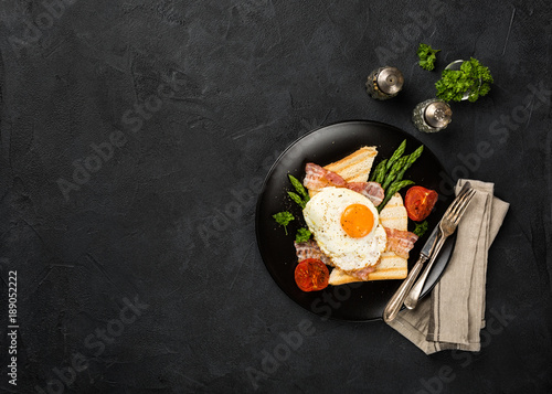 Overhead shot of breakfast or lunch with fried egg, bread toast, green asparagus, tomatoes and bacon on black plate with copy space. photo