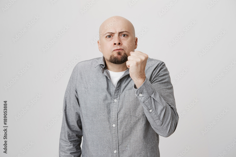 Portrait of serious male with beard looking angry holding his fist over white background. Bald man in casual clothes tells his teenage neighbor not to bother his cat unless he don't want troubles. Portrait of serious male with beard looking angry holding his fist over white background. Bald man in casual clothes tells his teenage neighbor not to bother his cat unless he don't want troubles.