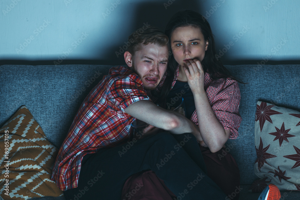 Naklejka premium scared guy with his girlfriend watching horror movie at night on sofa in home interior