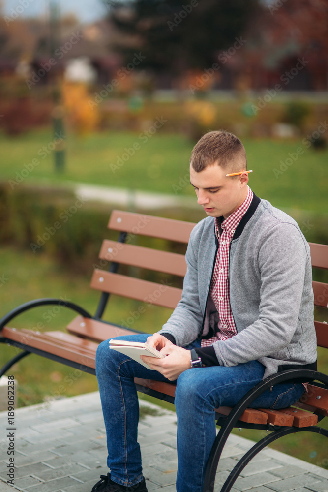 A cute student writes down his thoughts in his notebook using a pencil. Sits on bench A cute student writes down his thoughts in his notebook using a pencil. Sits on bench