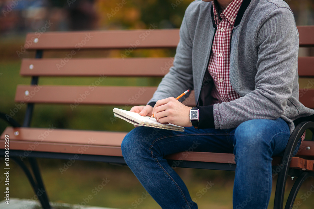 A cute student writes down his thoughts in his notebook using a pencil. Sits on bench A cute student writes down his thoughts in his notebook using a pencil. Sits on bench