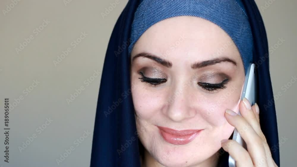 Portrait close-up of a beautiful Arab woman in a traditional headscarf hijab communicates on a mobile phone. A Muslim girl speaks on a white smartphone and smiles. Portrait close-up of a beautiful Arab woman in a traditional headscarf hijab communicates on a mobile phone. A Muslim girl speaks on a white smartphone and smiles.
