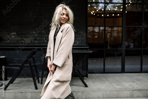 Portrait of beautiful young stylish blonde woman wearing beige coat and walking through the city streets. Trendy casual outfit. Street fashion. photo