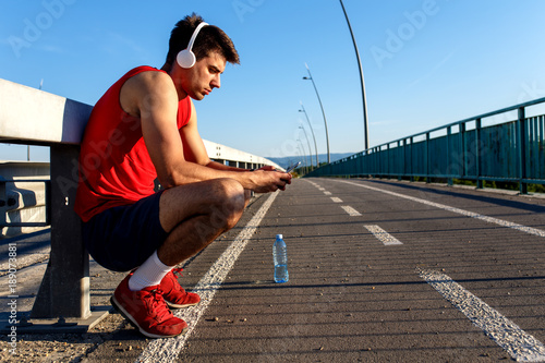 Fototapeta Naklejka Na Ścianę i Meble -  Young male runner with headphones looking at his mobile phone preparing for workout on bridge.