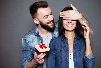 The gift will be a second. A young man gives his beloved a present and closes her eyes with a hand-rolled on gray background. Valentine's Day. Women's Day.