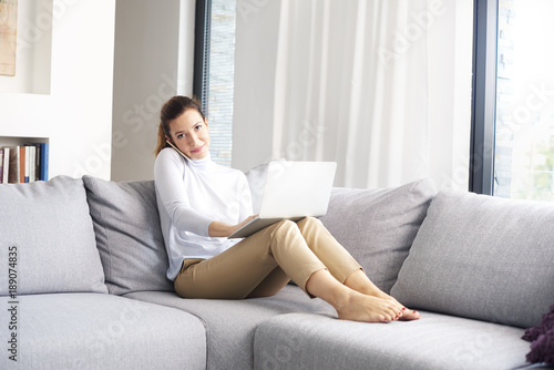 Managing her business from home. An attractive middle aged woman using her cell phone and making call while sitting on sofa with laptop and working from home office.