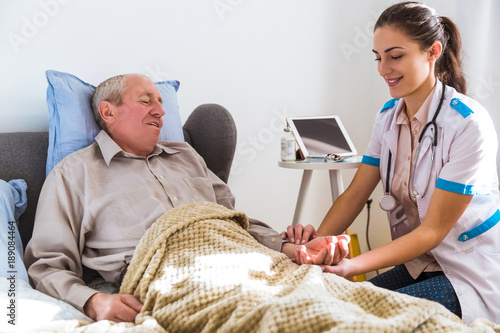 The young smiling doctor sitting near the old sick man and measuring his pulse at home