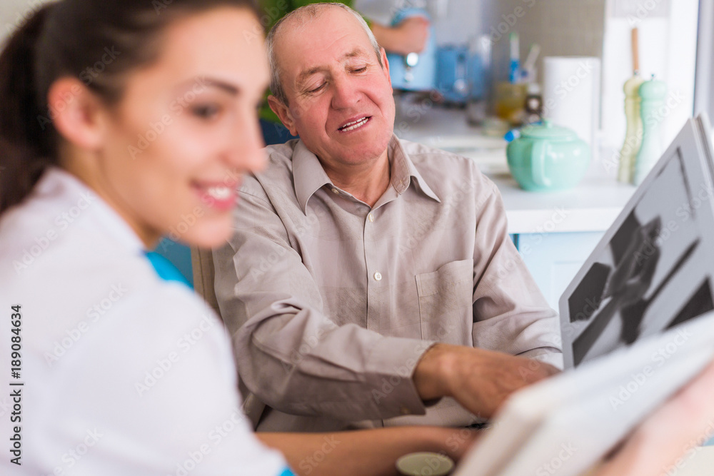 Obraz premium The old happy man sitting at the table with the young cheerful doctor and they reviewing photo album in the kitchen