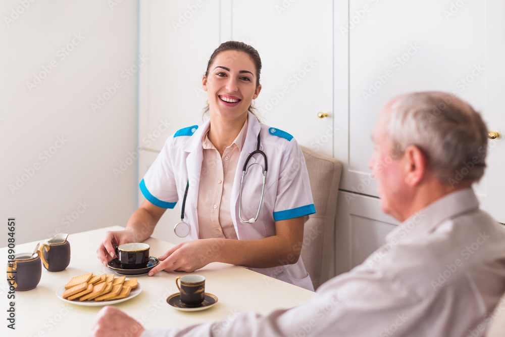 The young smiling doctor sitting at the table near ill old man and they drinking tea at home The young smiling doctor sitting at the table near ill old man and they drinking tea at home