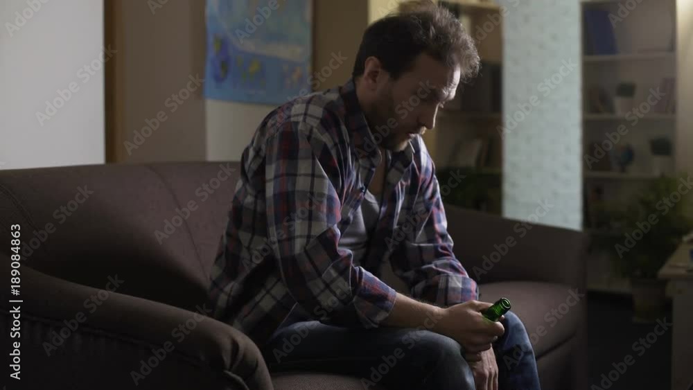 Drunken man sitting on sofa and looking at beer bottle, having hiccups, alcohol Drunken man sitting on sofa and looking at beer bottle, having hiccups, alcohol