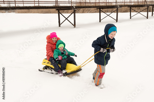Two boys and girl having fun sleigh ride during snowfall. Children sledding on snow. Siblings riding a sledge. Sport yellow sled in snowy winter park. Active fun for family vacation.