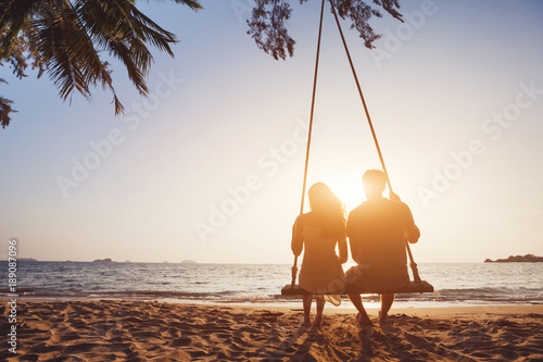 romantic couple in love sitting together on rope swing at sunset beach, silhouettes of young man and woman on holidays or honeymoon photo