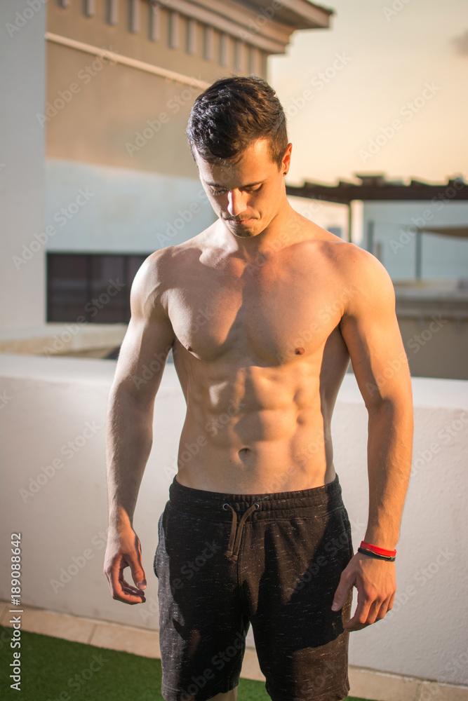 Bare-chested handsome muscular man in shorts standing outdoors and looking down. Bare-chested handsome muscular man in shorts standing outdoors and looking down.