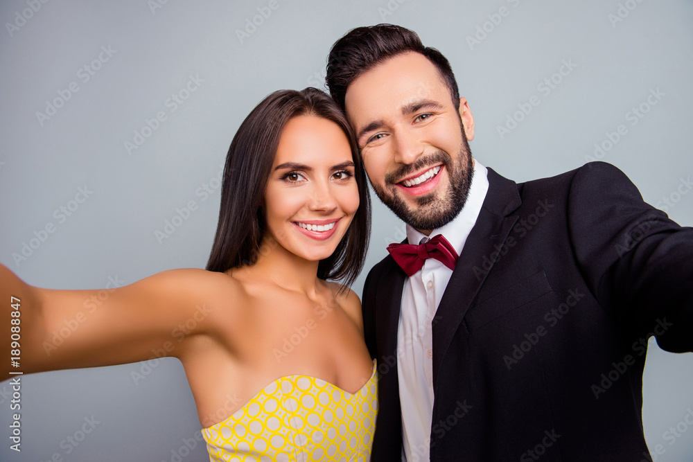 Lovely, cute, smiling, attractive, sexy couple, husband and wife in elegant  outfit, tux, dress with beaming smiles making selfie together in two hands  over grey background, 14 february foto de Stock
