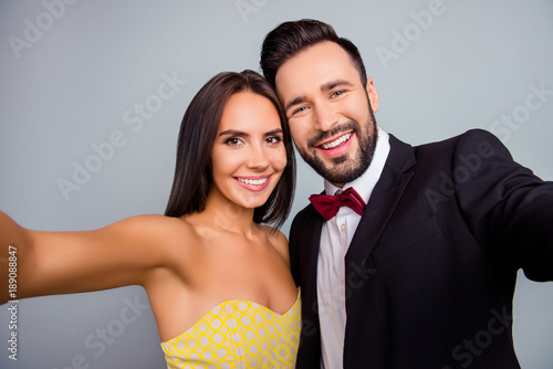 Lovely, cute, smiling, attractive, sexy couple, husband and wife in elegant outfit, tux, dress with beaming smiles making selfie together in two hands over grey background, 14 february