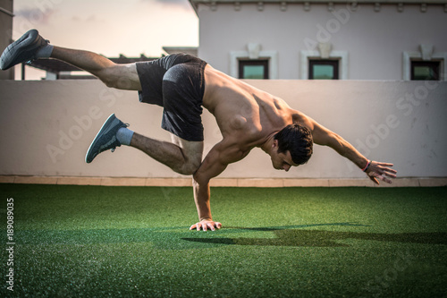 Fotografie Young athletic man balancing while doing a one hand push up and holding his body in the air outdoors