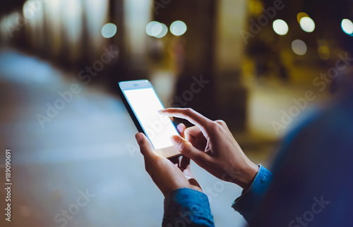 Woman pointing on blank screen smartphone on background bokeh light in night atmospheric city, hipster using in hands clean gadget mobile phone closeup, mockup street, online wifi internet concept