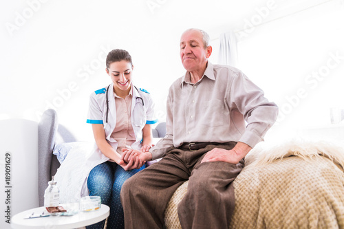 The old ill man sitting on a bed in the room at home and the young serious nurse sitting near him and measuring his pulse