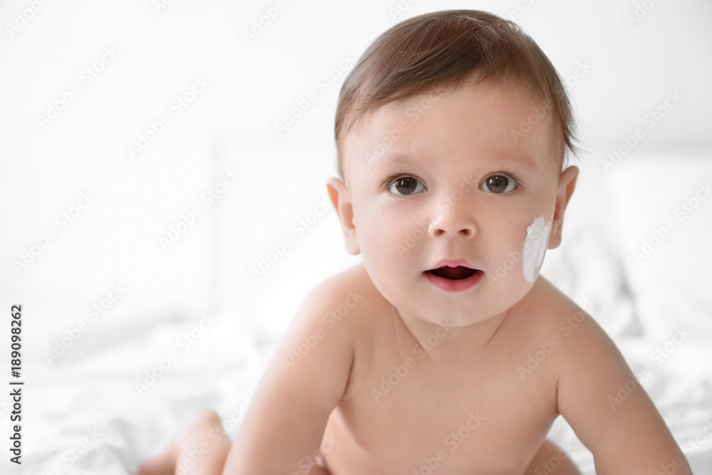 Cute baby with body cream on cheek against light background