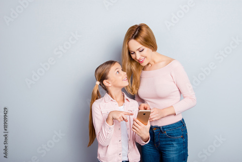 Attractive, nice, cute kid indicate with index finger on digital smart phone screen to her lovely mother, showing something interesting, together doing online shopping