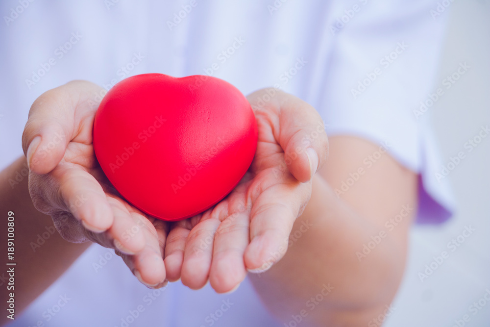 A nurse holding red heart toy. She is Left / right hand holding it. She is smile and good mood. The photo shows the principle of caring and good health. A nurse holding red heart toy. She is Left / right hand holding it. She is smile and good mood. The photo shows the principle of caring and good health.