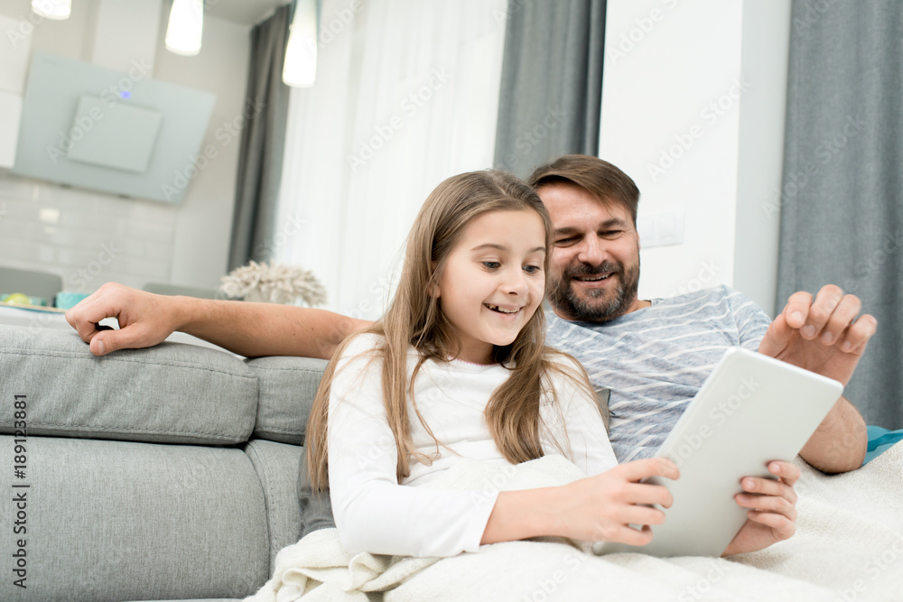 Bearded middle-aged man and his little daughter having fun together while sitting on cozy sofa and watching cartoons on digital tablet Bearded middle-aged man and his little daughter having fun together while sitting on cozy sofa and watching cartoons on digital tablet