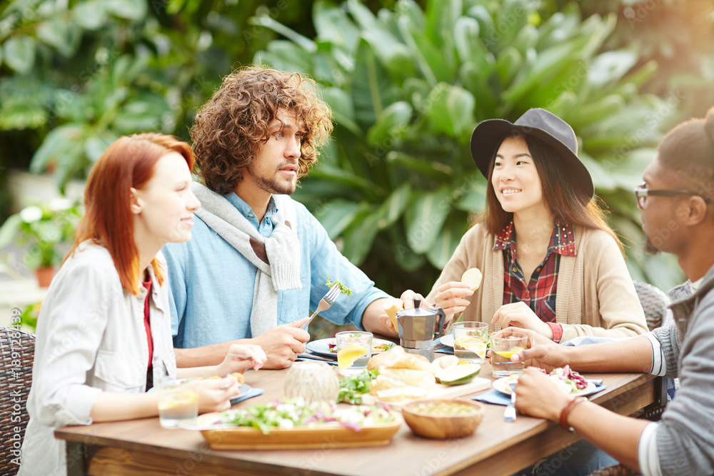 Young friends or two couples eating homemade food by table in the garden-room and discussing their plans Young friends or two couples eating homemade food by table in the garden-room and discussing their plans