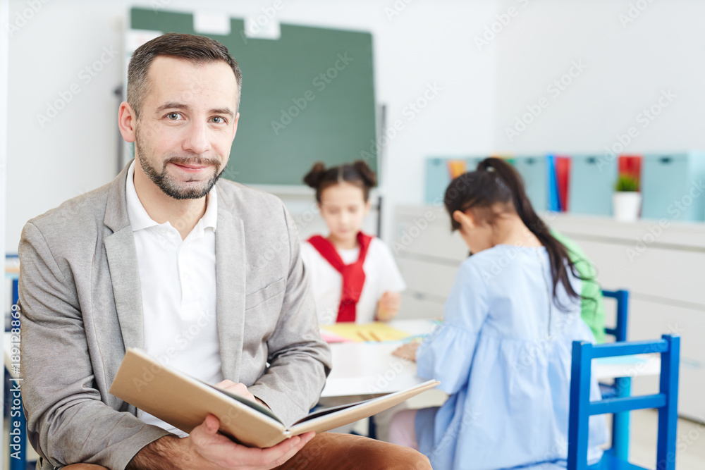 Mid-aged teacher with open book looking at camera on background of his pupils drawing by table Mid-aged teacher with open book looking at camera on background of his pupils drawing by table
