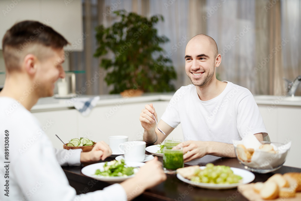 Happy young homosexual couple sitting by table in the kitchen and having breakfast