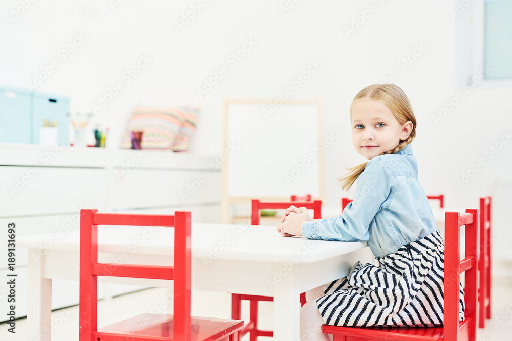 Cute little girl looking at camera while sitting by table on red chair in kindergarten Cute little girl looking at camera while sitting by table on red chair in kindergarten
