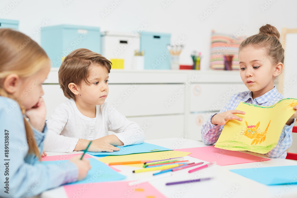 Cute elementary learner showing her picture drawn with crayons to her friends and explaining what it is Cute elementary learner showing her picture drawn with crayons to her friends and explaining what it is