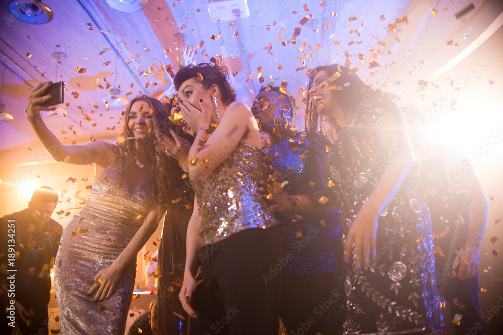 Group  of beautiful young women wearing glittering dresses dancing under golden confetti and taking selfies enjoying raving party in nightclub - obrazy, fototapety, plakaty 