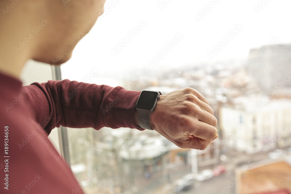 The man stands and is looking on his smart watches on his hand in front of window with view on city The man stands and is looking on his smart watches on his hand in front of window with view on city