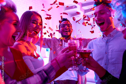 Group of happy friends enjoying holiday celebration at private house party raising glasses  and toasting under bursts of confetti