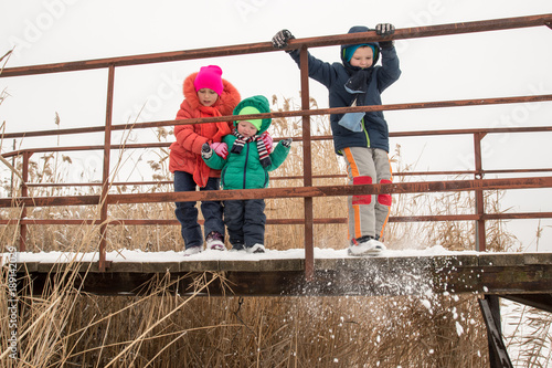 Cute little girl and boys in colorful winter clothes, playing on the bridge over a frozen river, outdoors during a snowfall. Active recreation in nature with children in winter