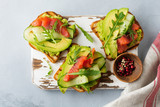 Open toast with trout, salmon, avocado, cucumber and arugula on wooden stand on gray concrete background. Selective focus. Top view. Copy space