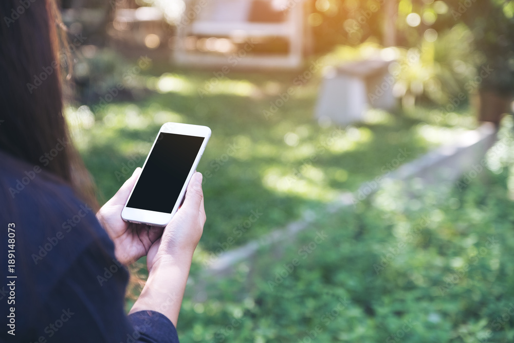 Mockup image of a woman holding and using white smart phone with blank black desktop screen in outdoor with green nature background