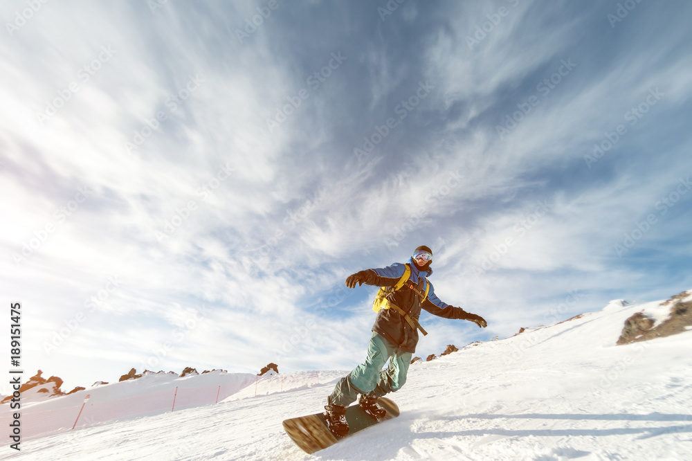 A snowboarder in a ski mask and a backpack is riding on a snow-covered slope leaving behind a snow powder against the blue sky and the setting sun