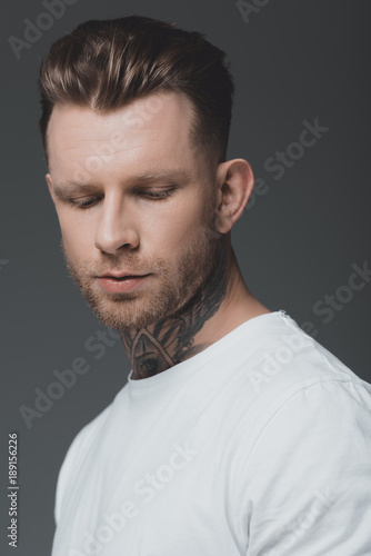 portrait of handsome stylish young tattooed man in white t-shirt looking down isolated on grey