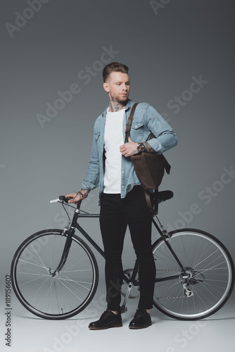 full length view of young tattooed man standing with bicycle and looking away on grey