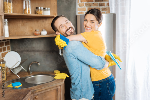 Funny cleaning. Cheerful energetic optimistic couple hugging while laughing and carrying cleaning cloth