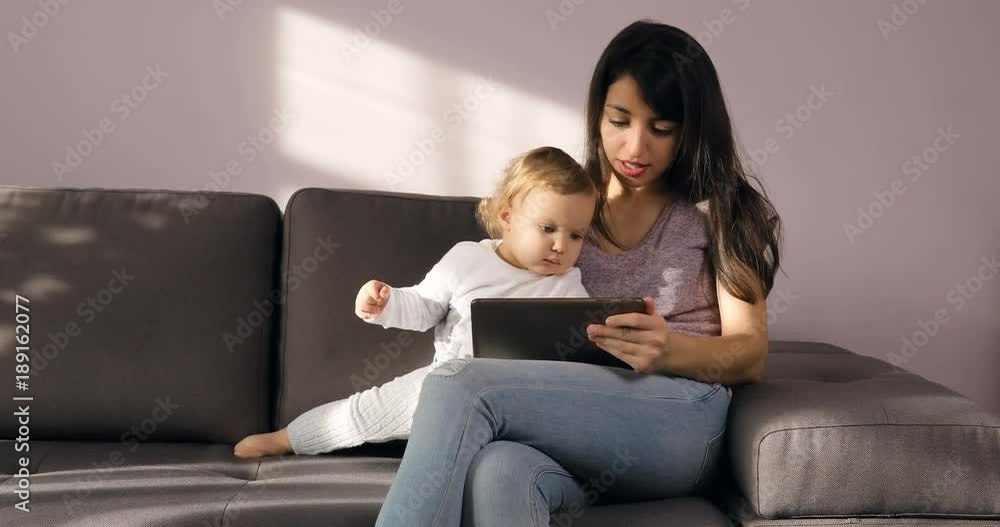 Brunette young women in casual clothes use tablet with little girl, show something on a stylish couch in sunny day Brunette young women in casual clothes use tablet with little girl, show something on a stylish couch in sunny day