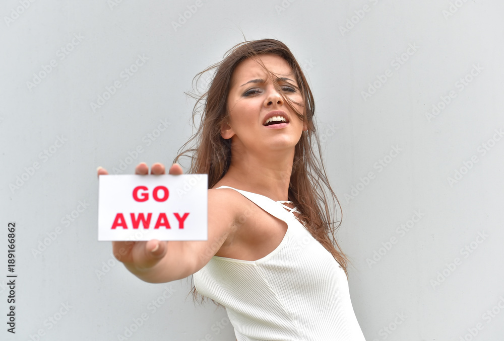A young girl stands in front of a grey cement wall. She holds out a white card with the words go away on it. She reveals a negative expression on her face. A young girl stands in front of a grey cement wall. She holds out a white card with the words go away on it. She reveals a negative expression on her face.