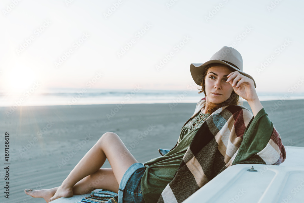 Charming young girl with hat, poncho, backpack with nice smile sitting on roof of recreational vehicle and having fun on the beach at sunset in warm weather. Boohoo style.