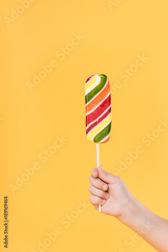 partial view of kid holding lollipop in hand isolated on yellow