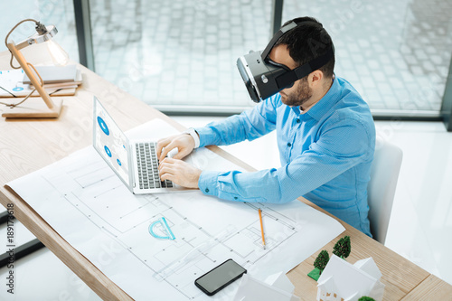 Engaged at work. The top view of a bristled young architect wearing a VR headset and working on a laptop while sitting at the work desk with a blueprint on it