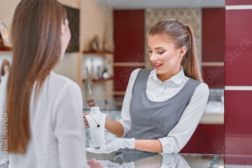 Valokuva Beautiful blonde jewelry worker presenting a costly necklace with a red stone inside to a woman with dark long hair and wearing a white blouse in a modern fashionable jewelry store