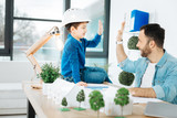 Great job. Cheerful little boy in a white hard hat sitting on the top of the table in his fathers office and giving him a high-five while the father working