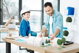 Clear explanation. Cheerful young architect explaining the basics of his work to his little son sitting on the top of the table and listening to him with a fond smile
