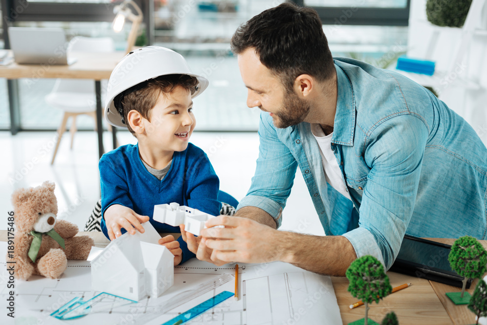 © Viacheslav Yakobchuk - New information. Pleasant young father and his little son in a white hard hat standing in the office near the table and discussing a 3D house model